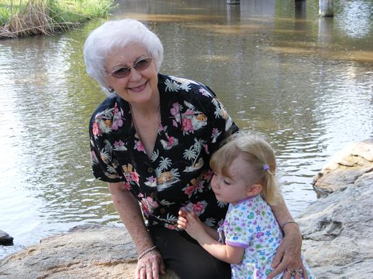 A cheerful elderly woman poses with a young girl near a riverbank, sharing a heartwarming moment.