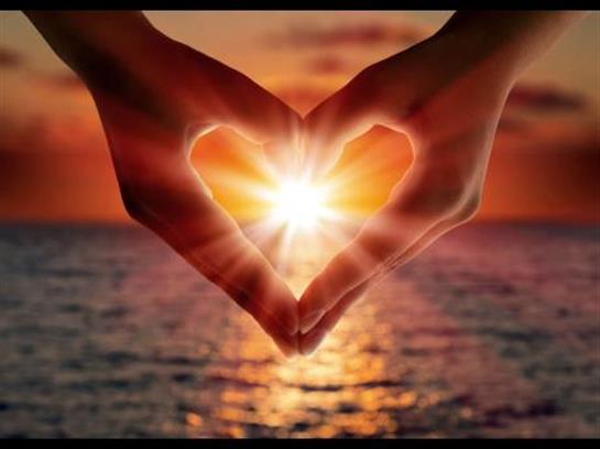 Two hands form a heart shape while a sunset reflects on the water, creating a romantic atmosphere.