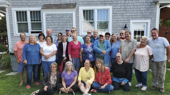 Friends of all ages gather happily outdoors, smiling and enjoying time together.