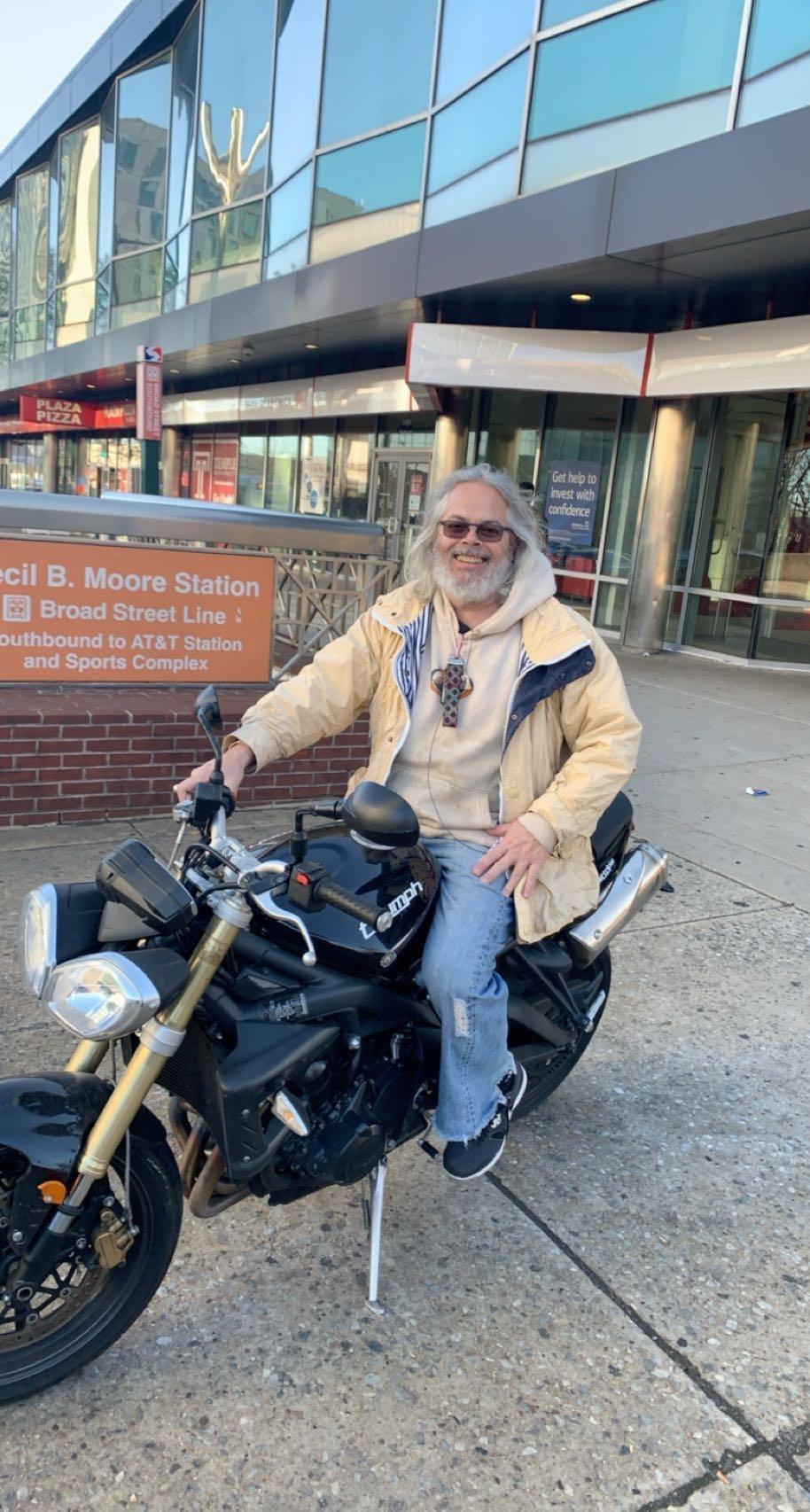 A man with long hair sits on a motorcycle at a train station in Philadelphia, radiating confidence.