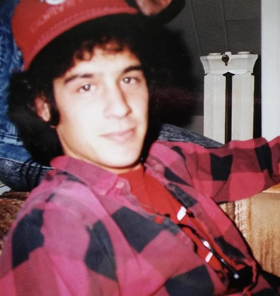 A young man sits comfortably on a couch, wearing a red hat and checkered shirt while smiling.