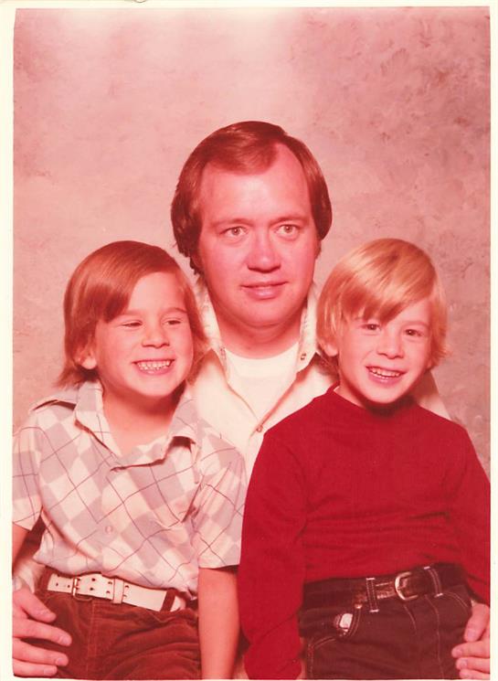 A father sits with his two sons, both smiling joyfully in a nostalgic setting.