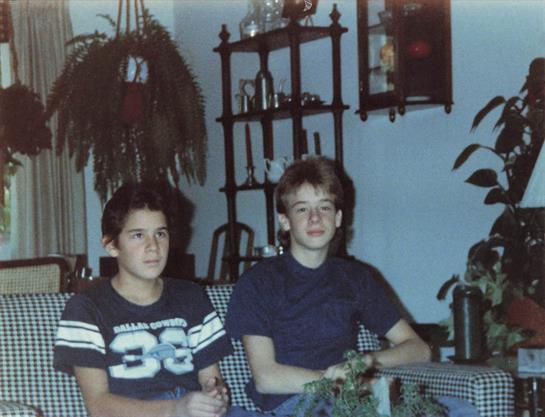 Two young boys relax together in a warmly decorated living space filled with plants.
