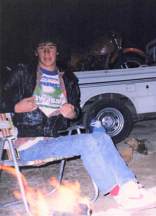 A young man sits in a camp chair, proudly displaying his shirt while surrounded by nature at night.