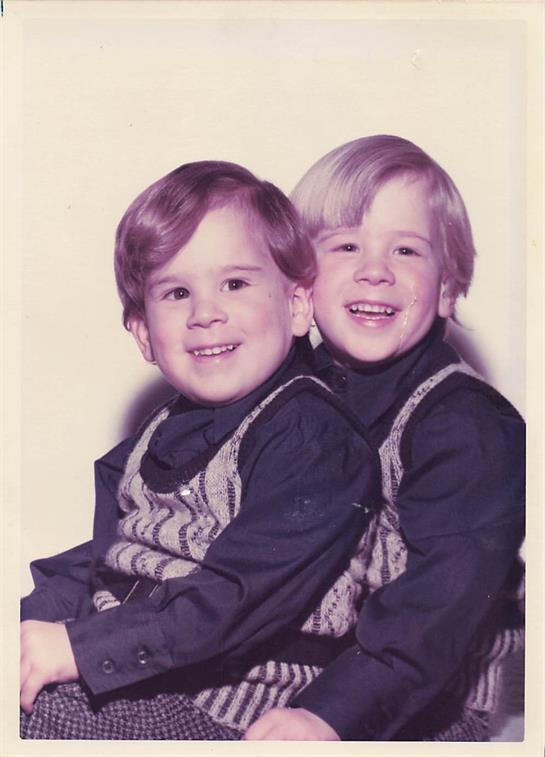 Two twin boys are sitting together, smiling happily, wearing similar sweaters and showcasing joy.