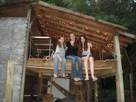 Three friends sit together on a treehouse deck surrounded by greenery and soft lighting.