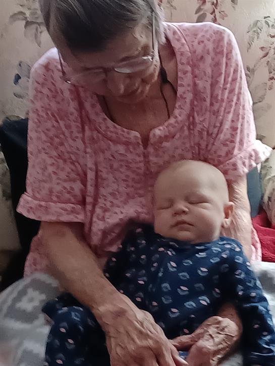 An elderly woman holds a sleeping baby on her lap in a cozy, peaceful living room.