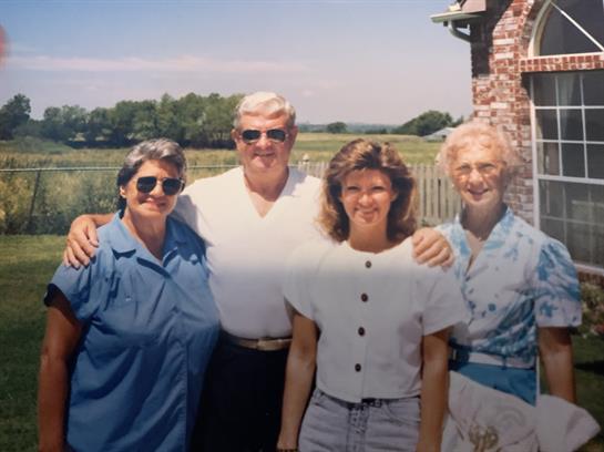 Four people stand closely together with smiles, enjoying a warm day in a rural landscape.