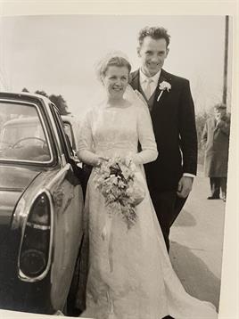 Newly married couple smiles as they leave vintage car, surrounded by friends and family in spring.