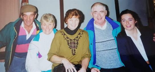 Three individuals are seated together, sharing smiles in a cozy indoor environment, dressed warmly.