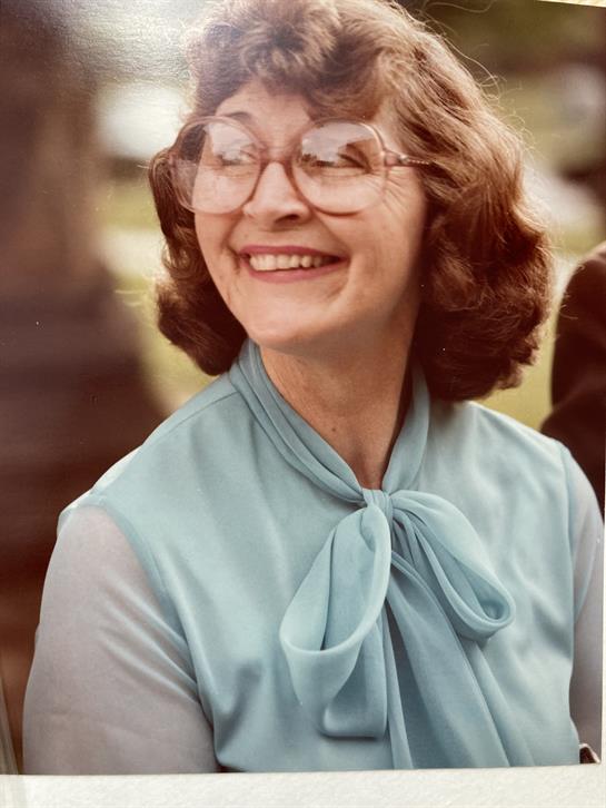 A woman with glasses and curly hair enjoys a sunny day at a park gathering, smiling brightly.