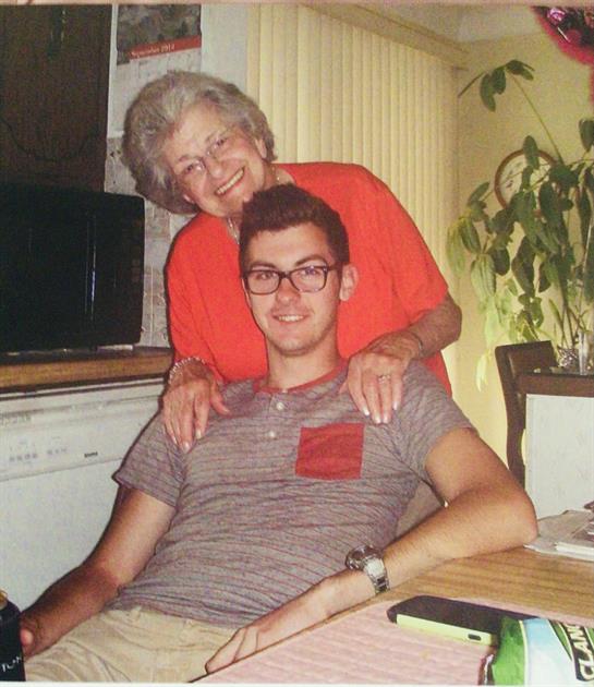 A young man sits at a kitchen table while an elderly woman hugs him from behind, both smiling.