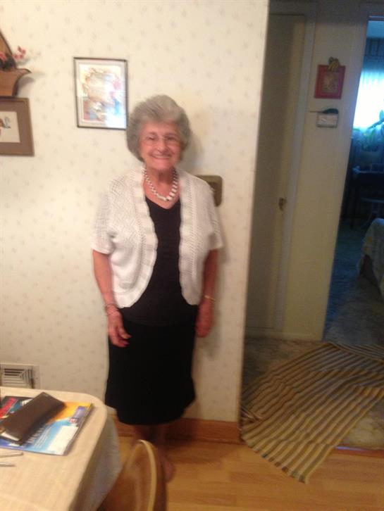 An elderly woman poses with a warm smile in her living room, surrounded by personal items.