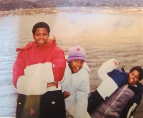 Kids smiling and posing near a serene body of water with mountains in the background during winter.