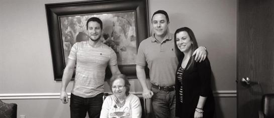 Group of four adults smiling together with an elderly woman seated, celebrating a special occasion.