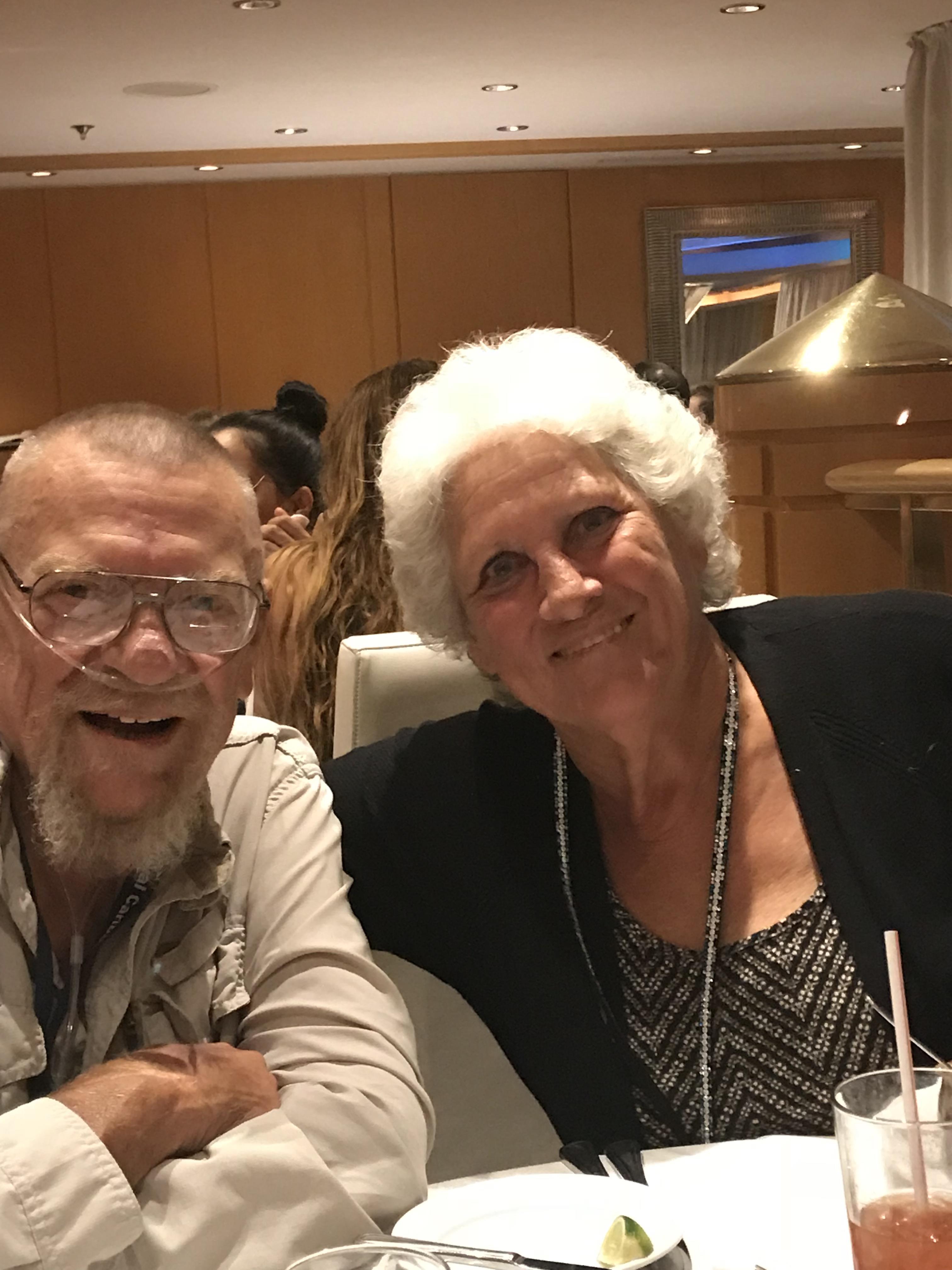 Two senior adults smile joyfully while dining out at a restaurant surrounded by family.