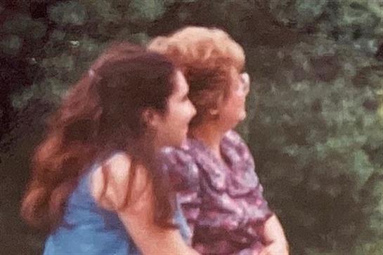 Two women are sitting side by side, smiling and engaging in a lively conversation outdoors.