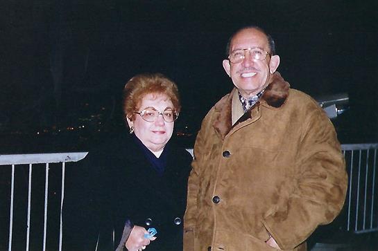 A couple stands together, smiling warmly, dressed for a chilly evening outdoors.