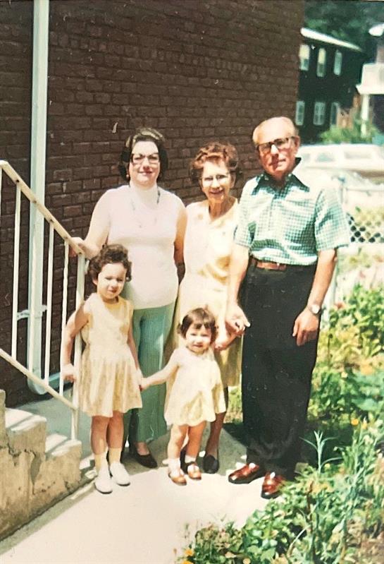 Three generations enjoy a warm day together outside a house, smiling and holding hands.