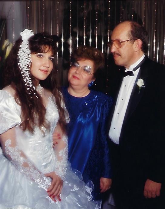 Bride stands happily beside her parents at a wedding, dressed in a beautiful gown with lace details.