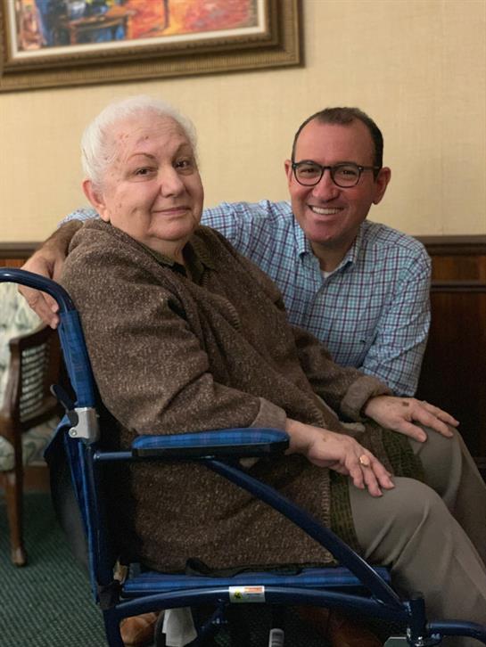 Elderly woman in wheelchair smiles while sitting beside a man in a casual checkered shirt.