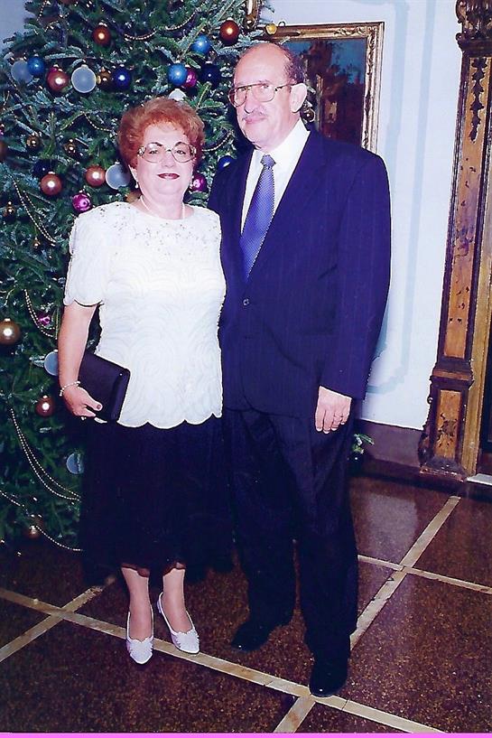 Couple stands together smiling in formal attire near a vibrant Christmas tree with ornaments.