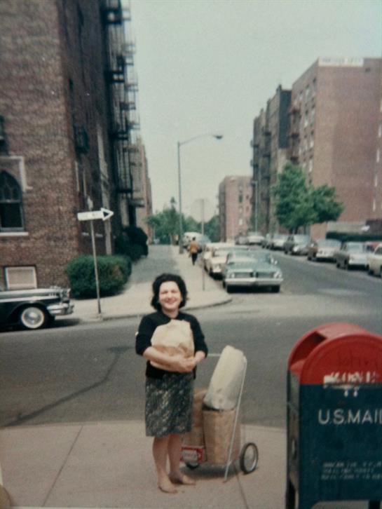 A woman stands on a city street with groceries, cars lined up in the background.