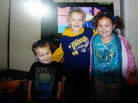 Three children are smiling and standing together in a living room, enjoying a fun moment.