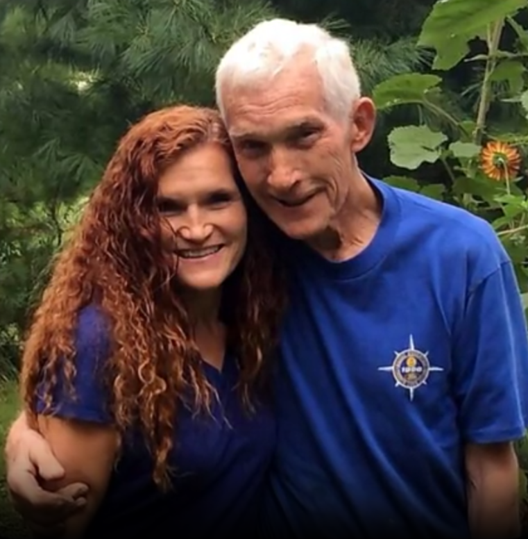 A joyful elderly man and a young woman smile in a bright garden of greenery and sunflowers.