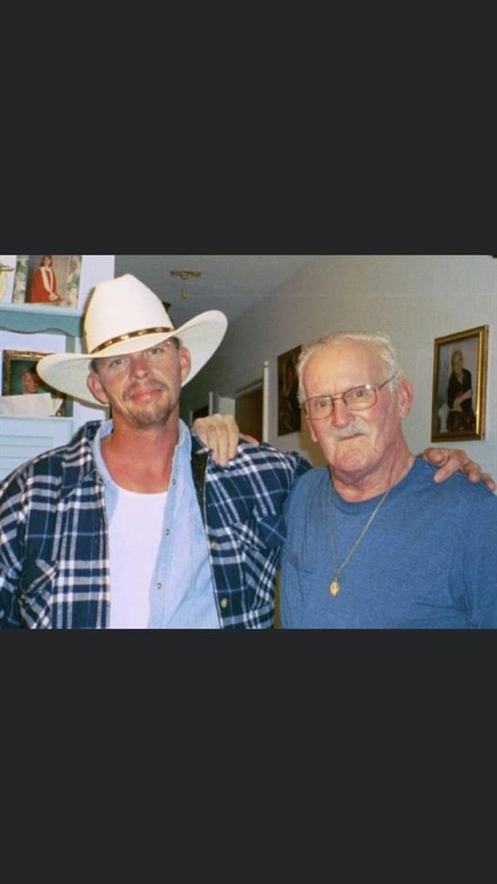 Two men stand closely, sharing smiles in a cozy interior adorned with family memories.