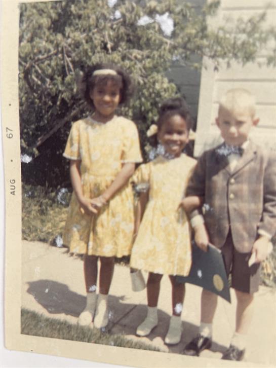 Three children wearing bright dresses and a suit stand together, smiling in a garden.