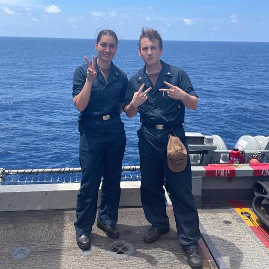 Two sailors smile and pose on the ship's deck under a bright, sunny sky.