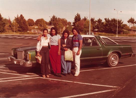 Four friends pose together in a parking lot with a vintage car during the 1970s.