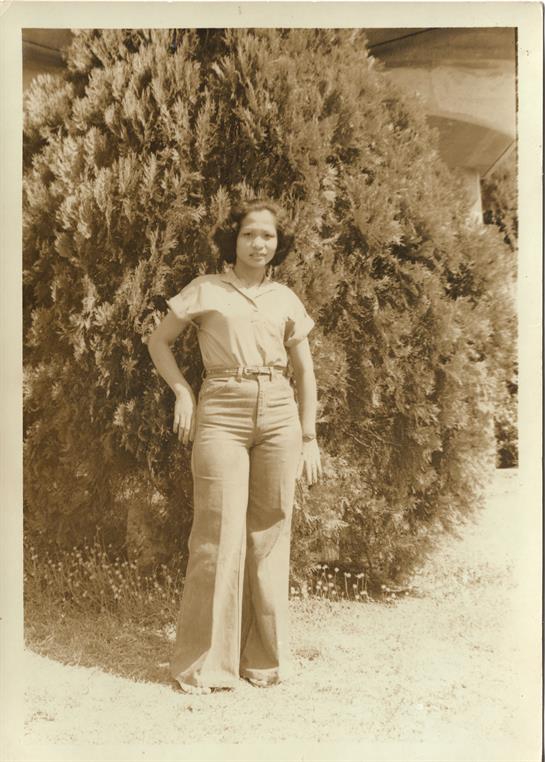 A woman poses in fashionable attire near dense greenery, exuding a sense of confidence and charm.