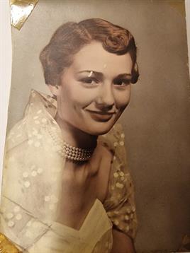 A woman poses gracefully with a warm smile, dressed in elegant vintage attire and jewelry.