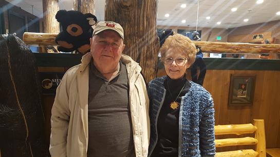 Elderly couple stands together, smiling and dressed casually, enjoying a social gathering indoors.