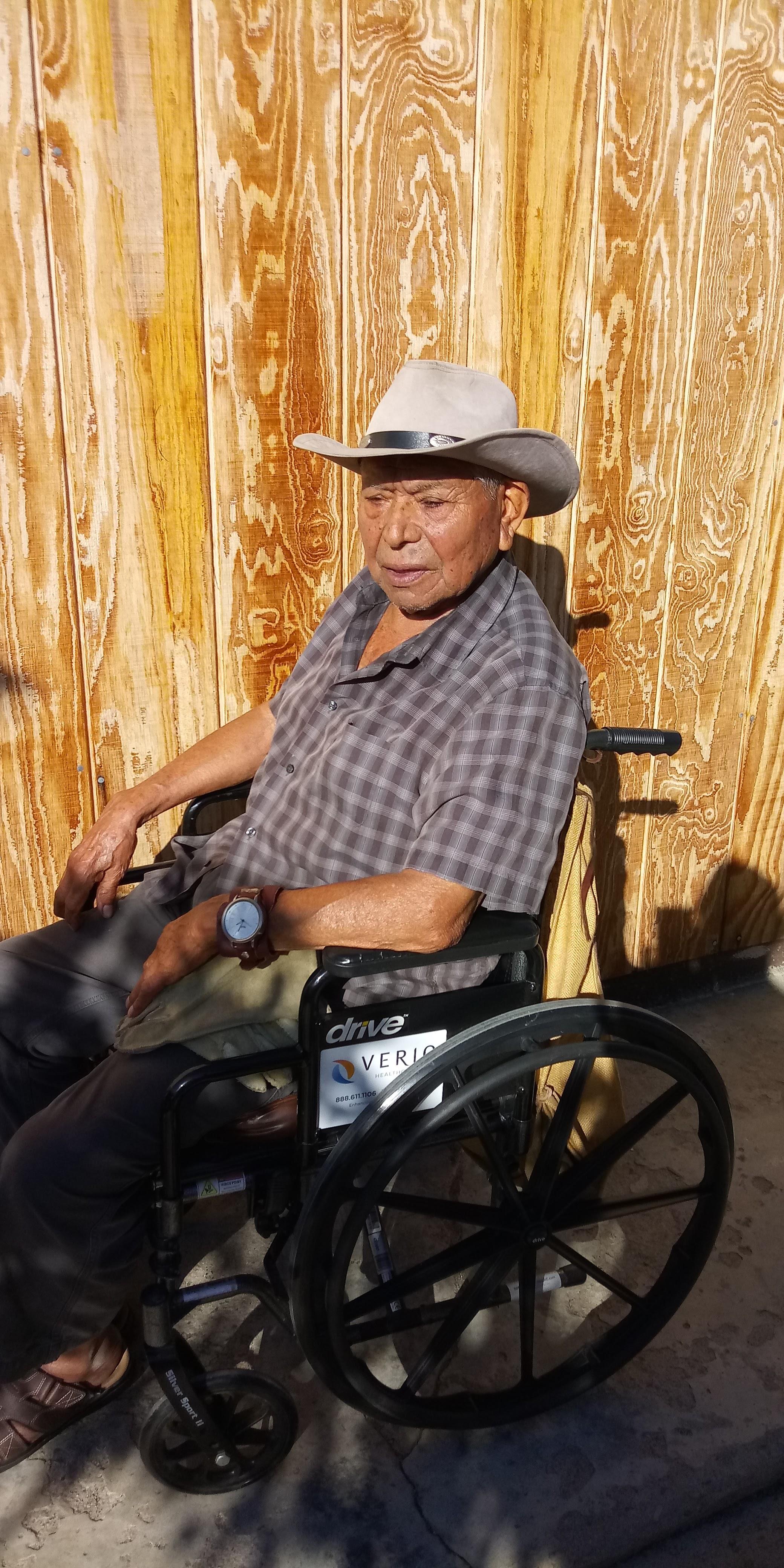 Older gentleman in a wheelchair sits comfortably under the sun, leaning back against a wooden wall.