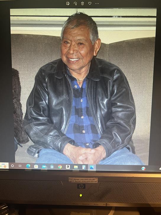 An elderly man in a leather jacket smiles warmly from a cozy living room couch.