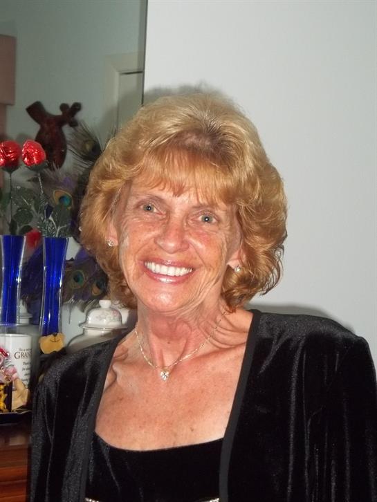 A cheerful middle-aged woman with curly hair smiles brightly in a cozy room decorated with flowers.