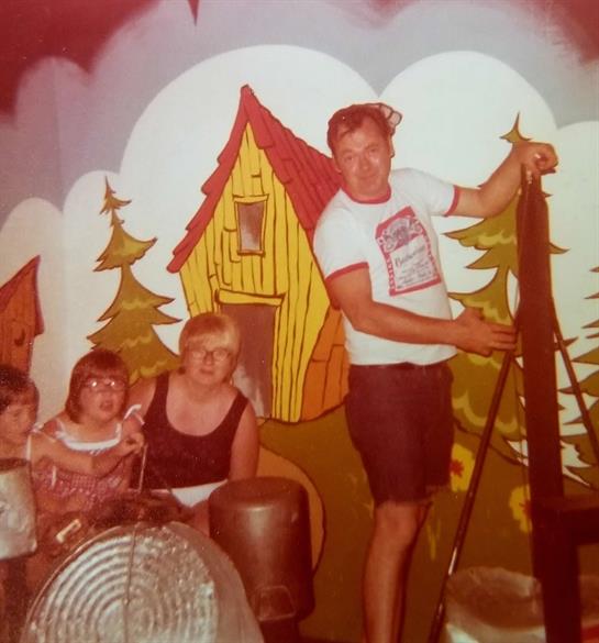 A family poses joyfully beside a colorful mural of a house and trees on a sunny day.