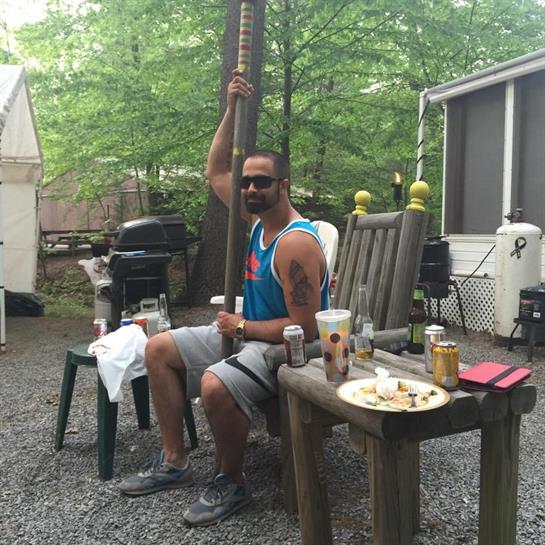 A man sits at a rustic table in a backyard, enjoying drinks and food during a sunny day.
