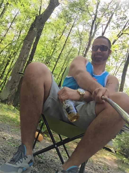 A man sits comfortably in a chair in the forest, taking a selfie while enjoying a beverage.