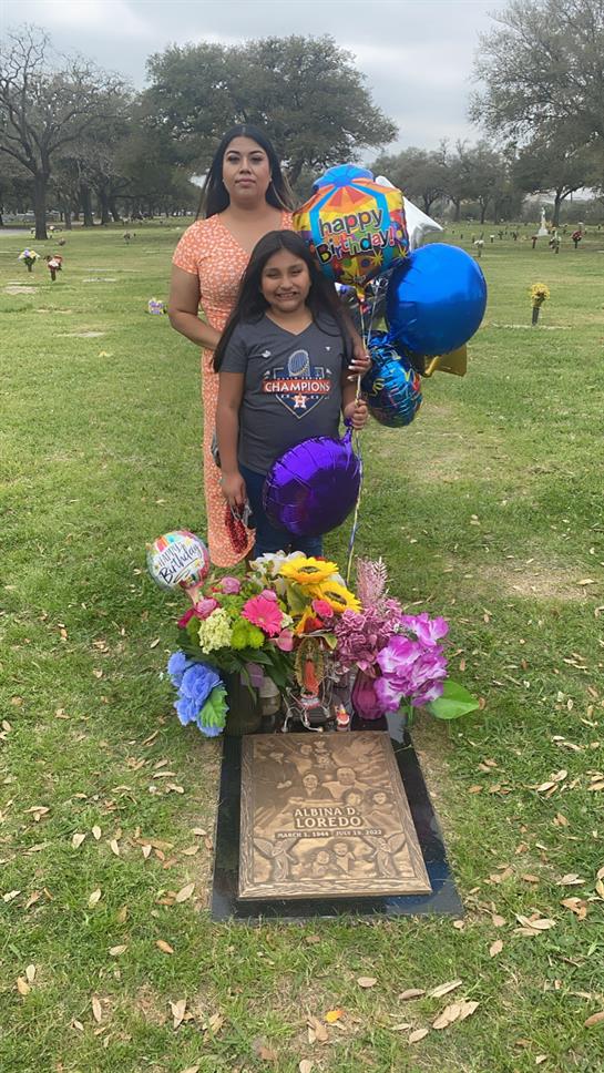 A woman and a girl stand at a grave, honoring a loved one's memory with flowers and balloons.