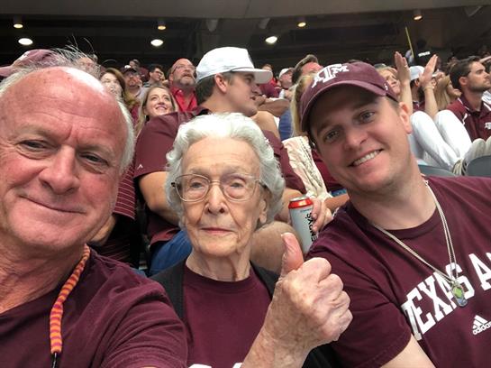 Three family members smile and celebrate at a lively football game in Texas with maroon shirts.