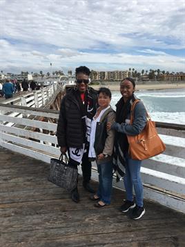 Three friends smile on a pier with ocean waves and a cloudy sky behind them.