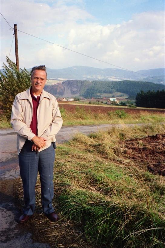A man poses confidently in a rural area, with scenic mountains and fields in the background.