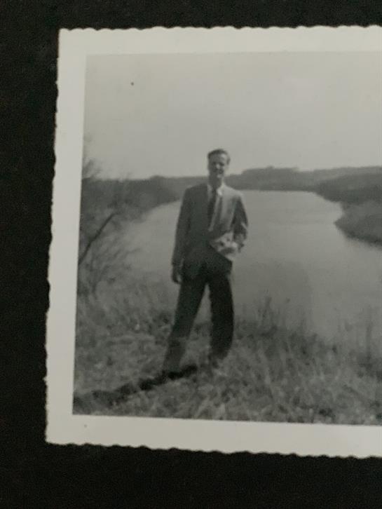 A man in a suit stands confidently on a riverbank, surrounded by lush grass and calm water.
