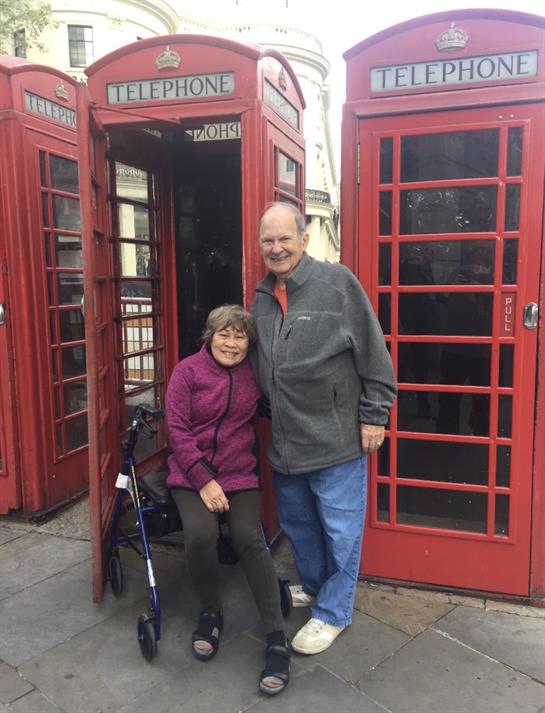 A couple poses happily together outside two vintage red telephone booths in a bustling city.