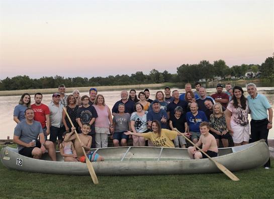 A large group gathers at the lake, smiling and posing together in a canoe during sunset.