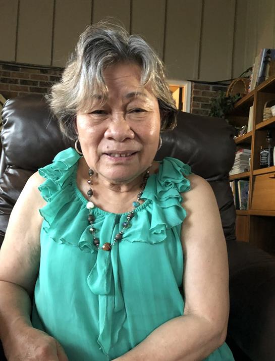 A senior woman in a turquoise top smiles while seated comfortably in her living room.